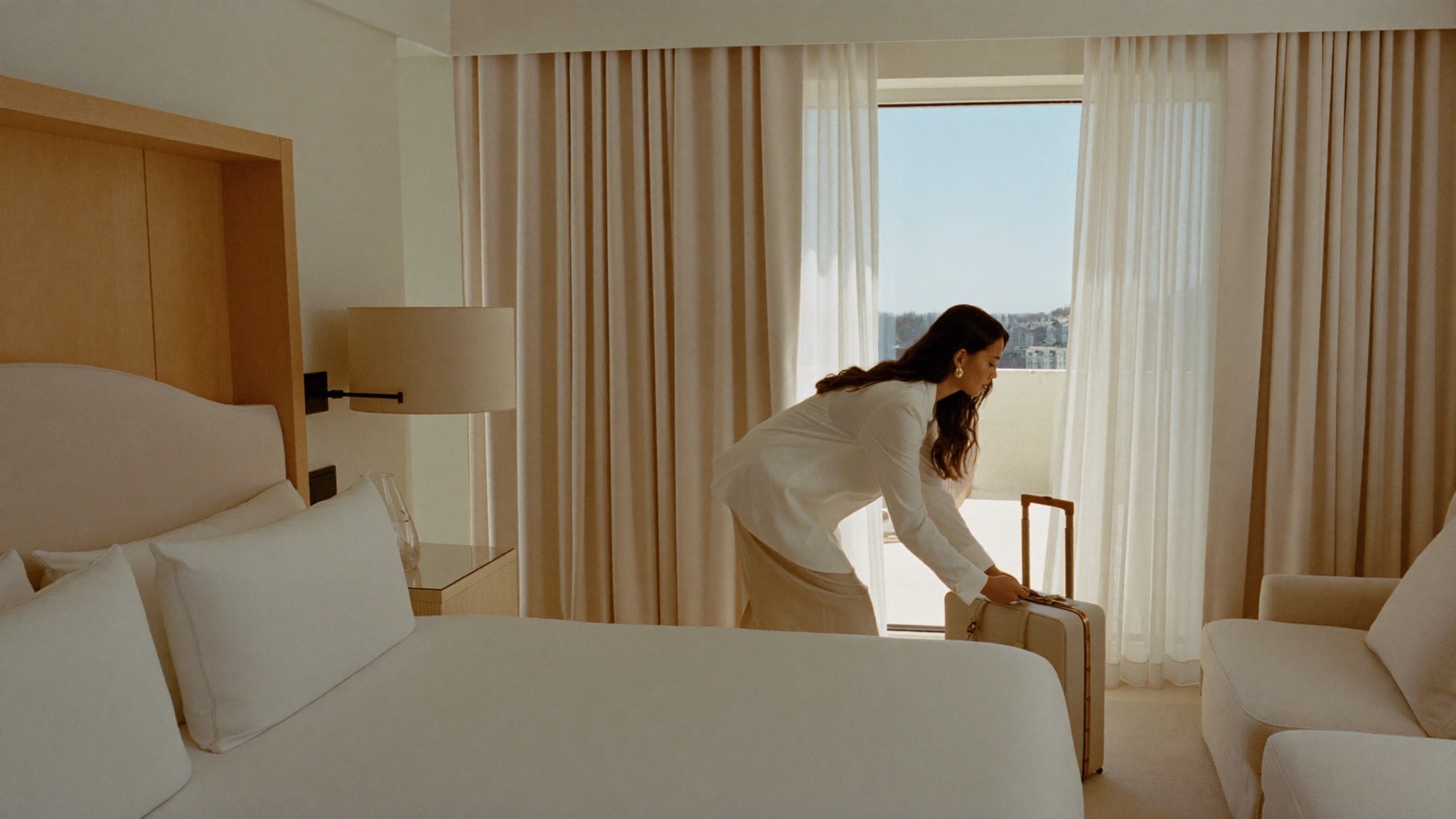 A woman arriving at a calm, light-filled recovery-stay suite, setting down her travel case